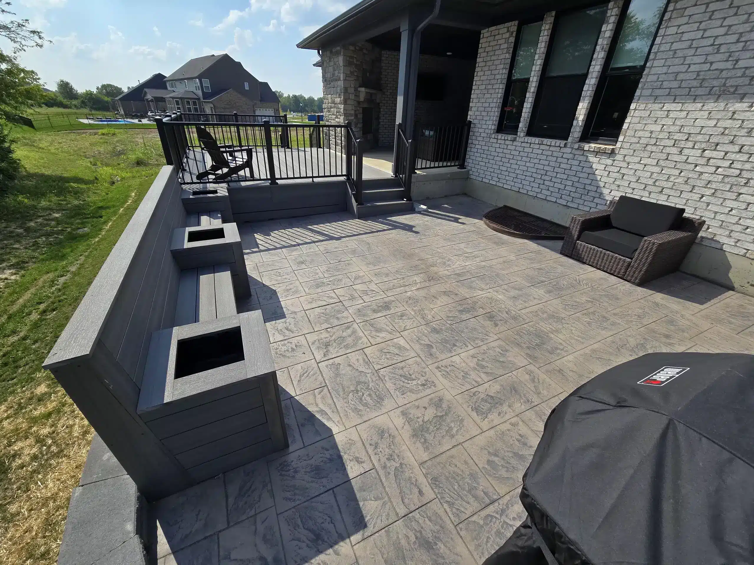 An outdoor patio area with gray-toned paving stones and a raised deck. The patio features an outdoor seating area with a dark wicker chair and a grill covered with a black cover. Along one side of the patio, a built-in bench features two square planters.
