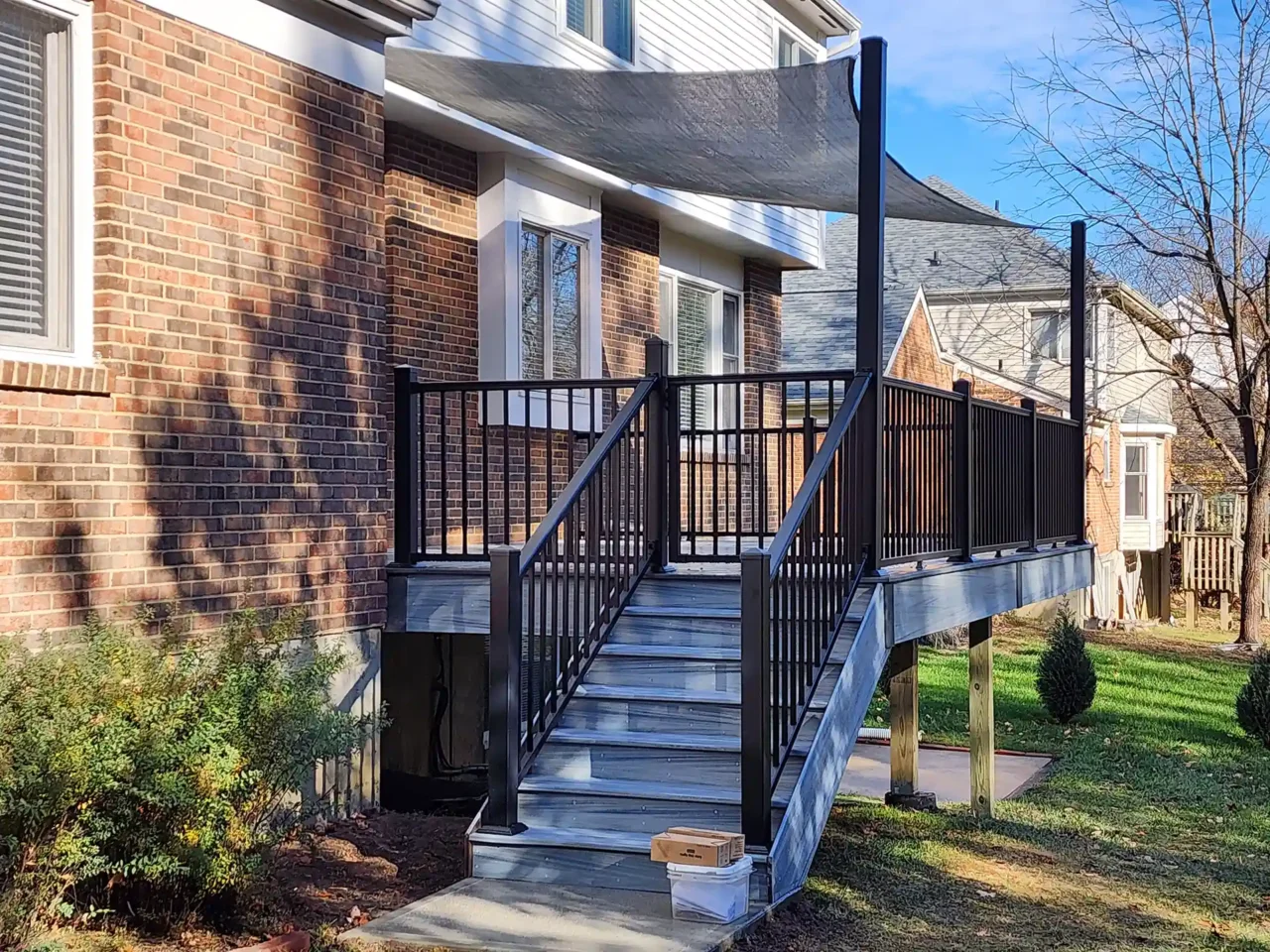 A raised deck with black metal railings and gray composite steps, leading down to yard, with a fabric sunshade above the deck, and a brick house in the background.