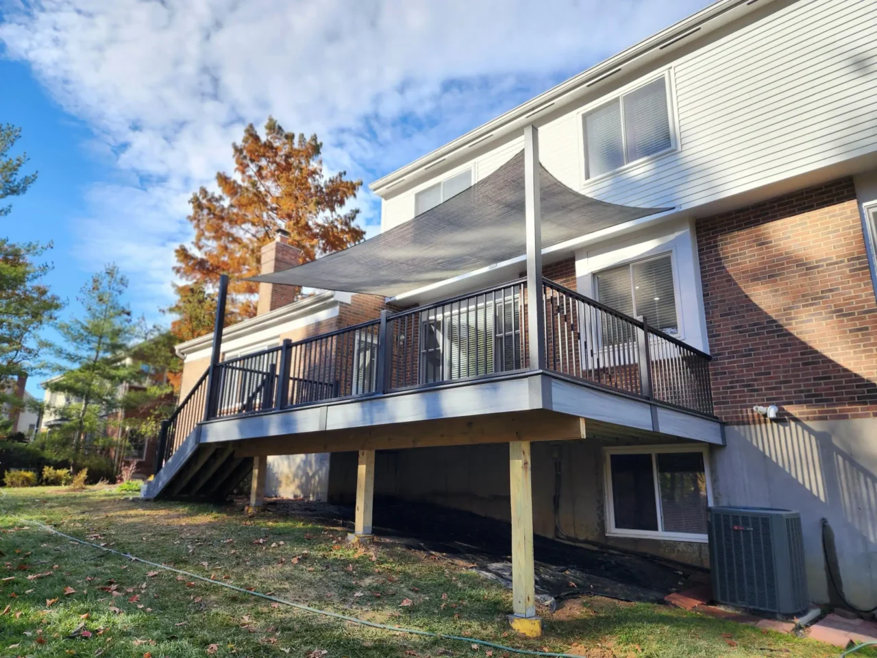 View from the side showing a raised deck with black railing, a staircase, and a gray sun sail above. The deck is attached to a brick house with a grassy yard.