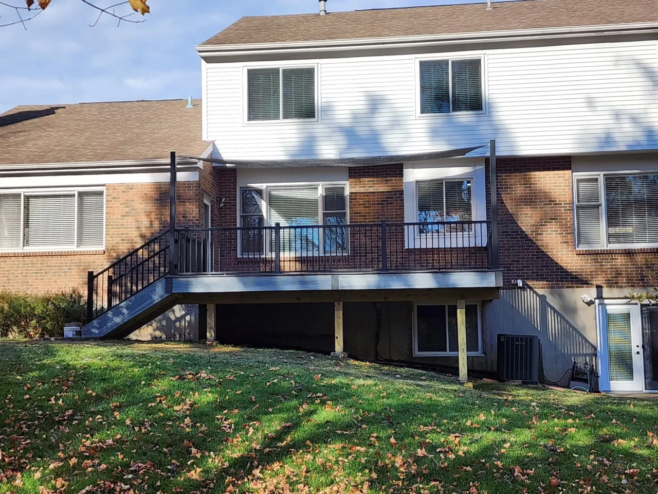 A raised deck with black railing and a staircase, attached to a brick house. The area underneath the deck is visible, with a grassy yard in front.