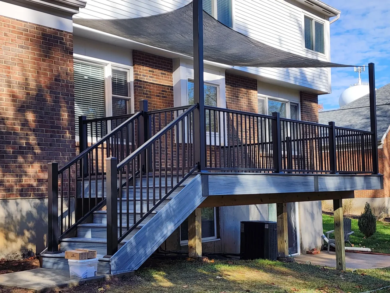 Side view of a raised deck with black railing, a staircase, and a gray sun sail above. The deck is attached to a brick house.