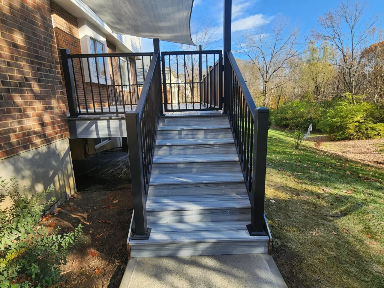 A staircase with black railing and gray composite steps, leading up to a raised deck attached to a brick house.