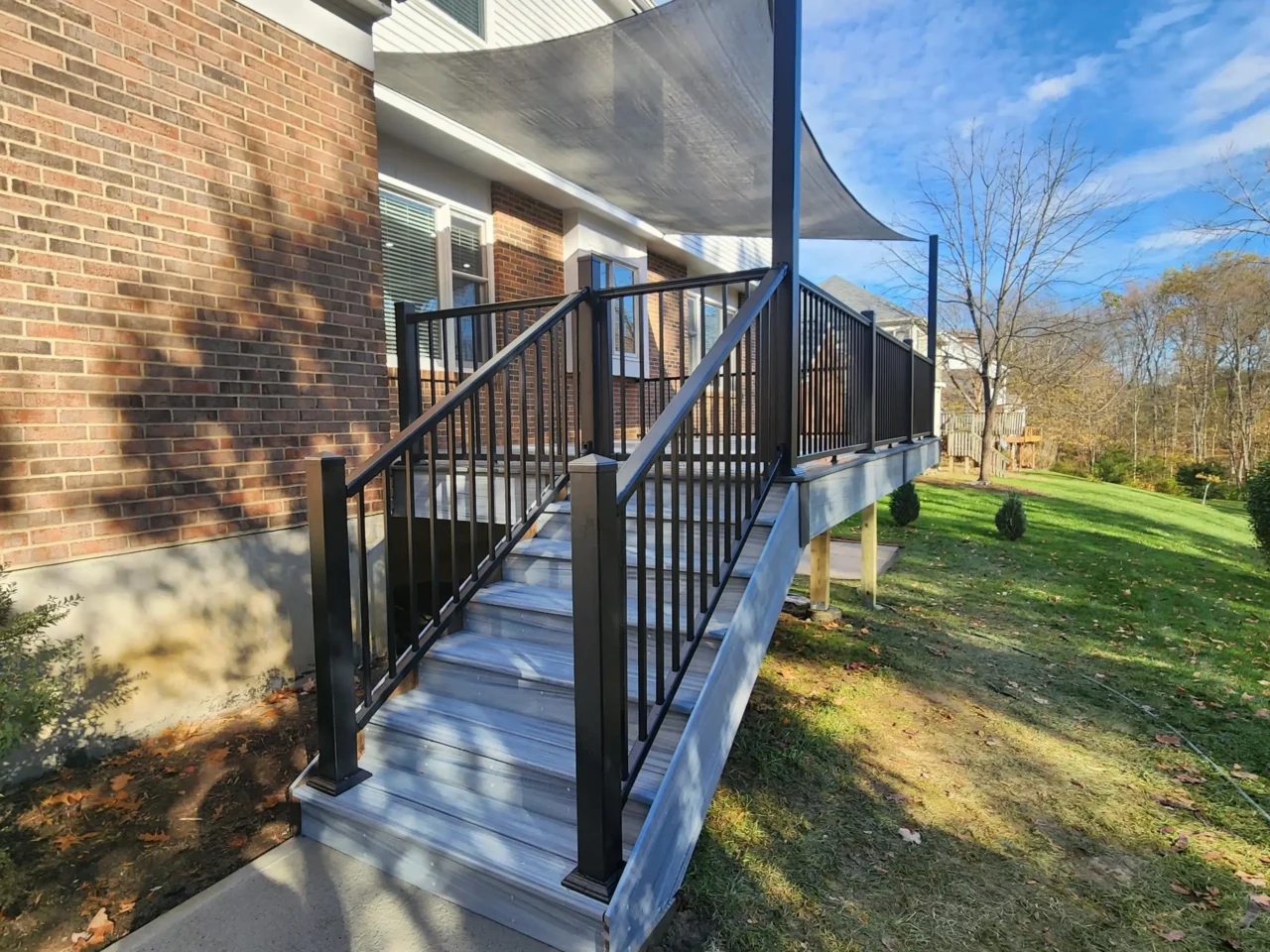 View from the front-left of a raised deck with black railing, a staircase, and a gray sun sail above. The deck is attached to a brick house.