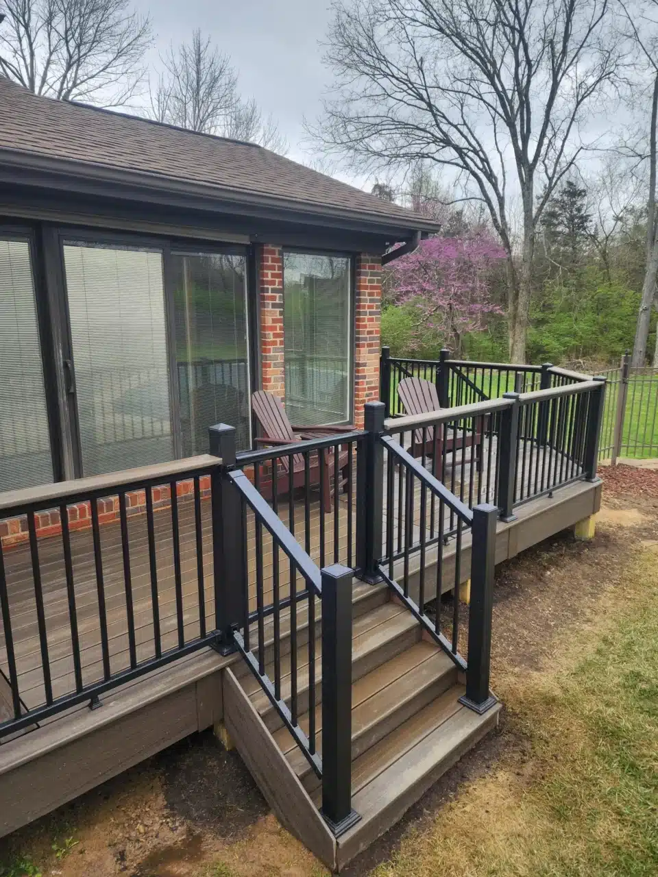 Side view of a raised deck with black railing and a staircase leading down to the yard. The deck is attached to a brick house with trees.
