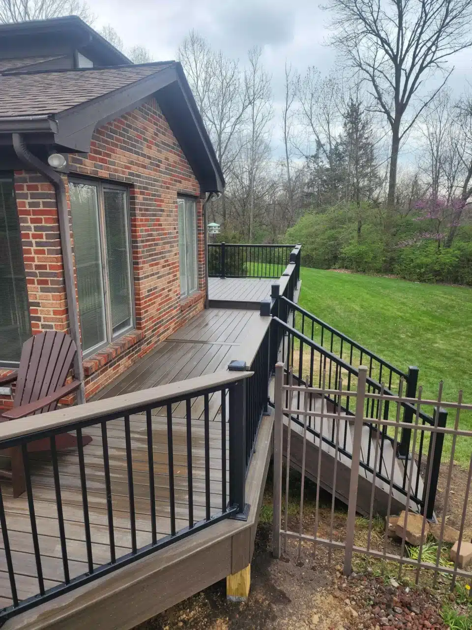 View from the side of a raised deck with black railing, featuring a staircase leading down to the yard. The deck is attached to a brick house.