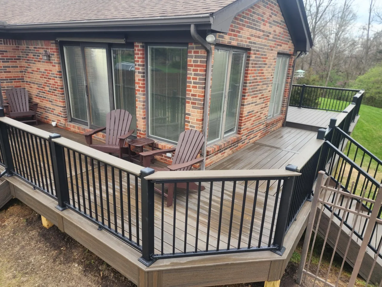 Overhead view of a raised deck with black railing, attached to a brick house. Two chairs are visible on the deck, and the staircase leads to the yard.