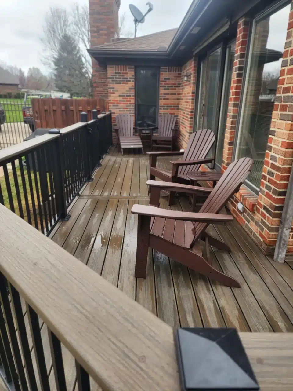 Close-up view of a raised deck with black railing and wooden decking. Two Adirondack chairs are placed on the deck, with a brick house visible in the background.