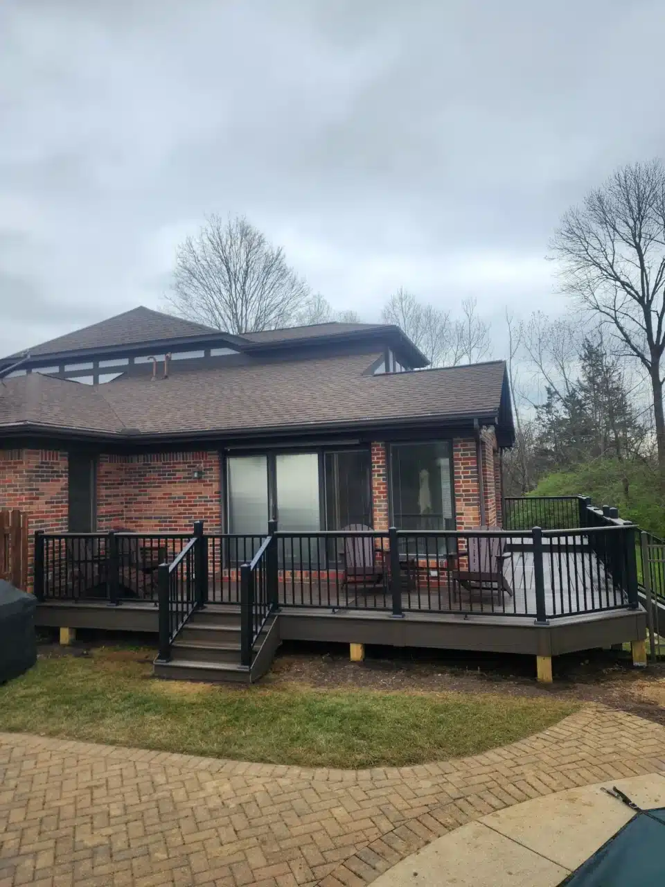 View from the front-right of a raised deck with black railing, attached to a brick house. The staircase leads to a paved area