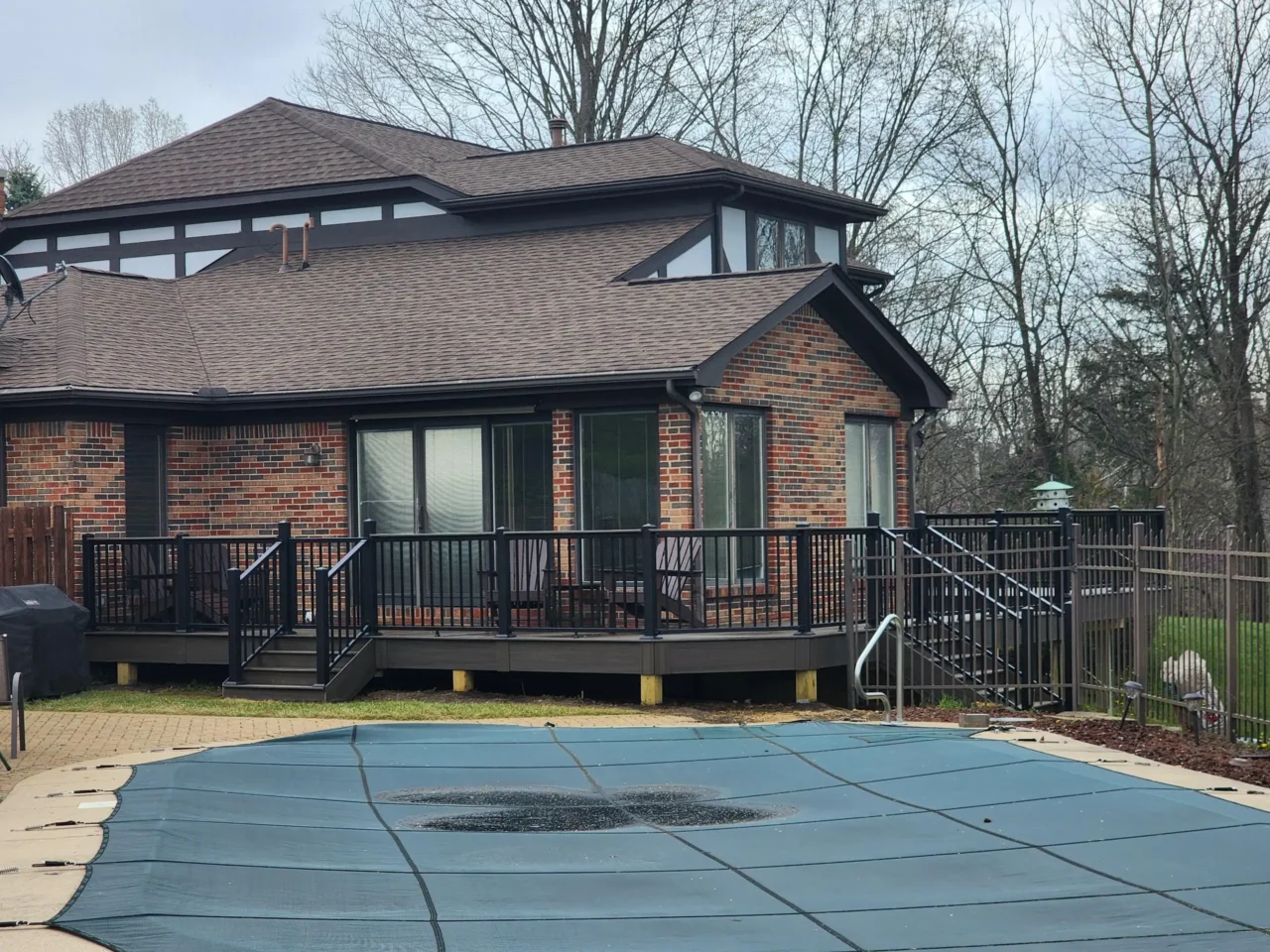 A raised deck with black railing, attached to a brick house. A pool cover is visible in the foreground, with a fenced yard and trees in the background.