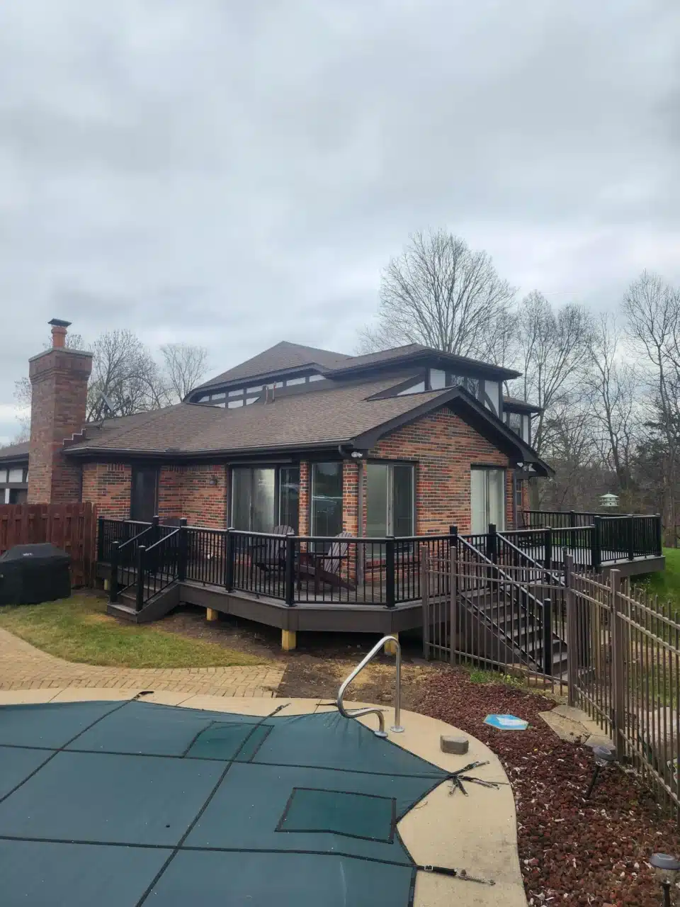 Side view of a raised deck with black railing, attached to a brick house. A pool cover is visible in the foreground, with a fenced yard and trees in the background.