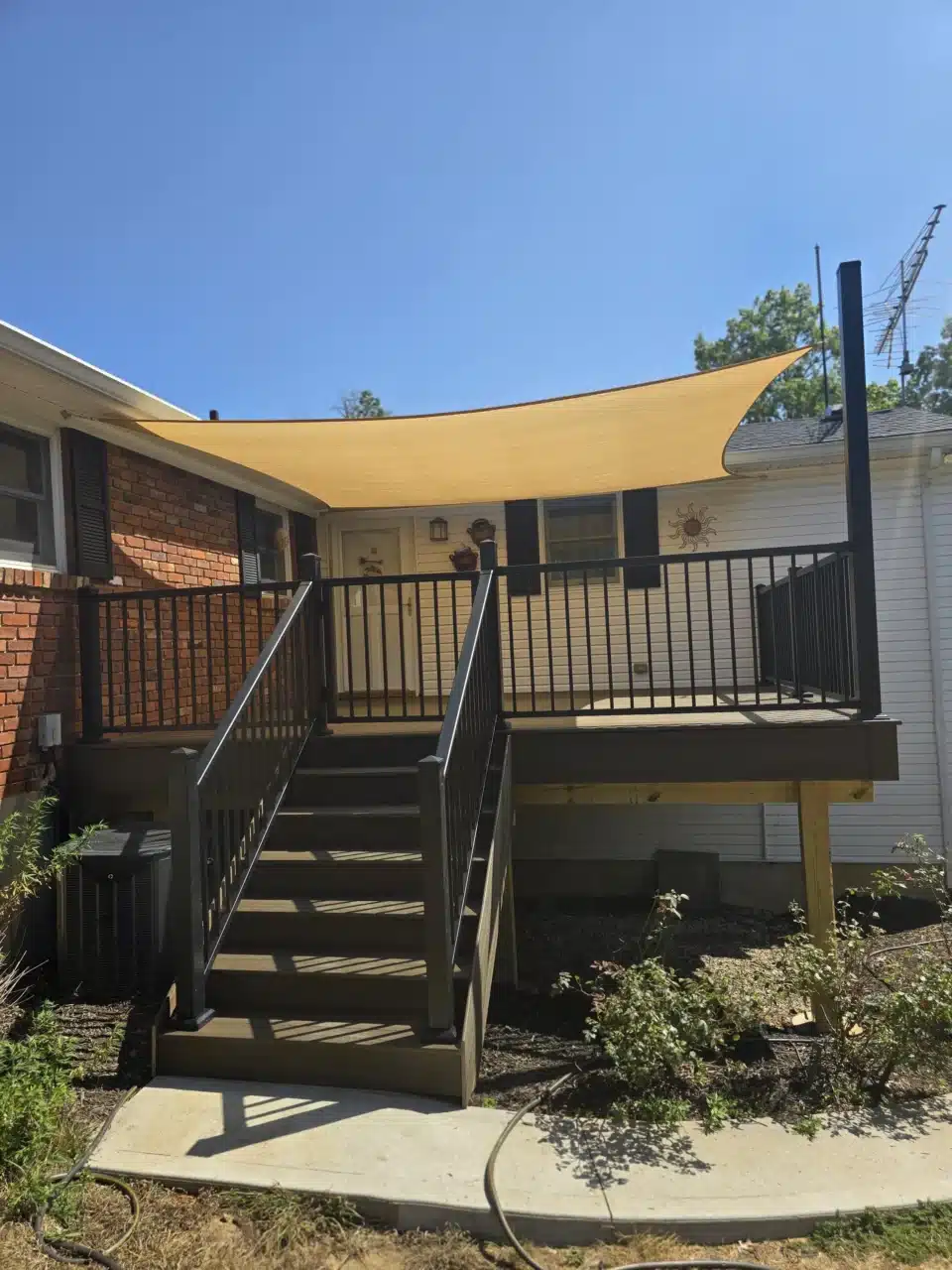 Front view of the elevated deck with black aluminum railing and a staircase. A shade sail extends above the deck, providing coverage.