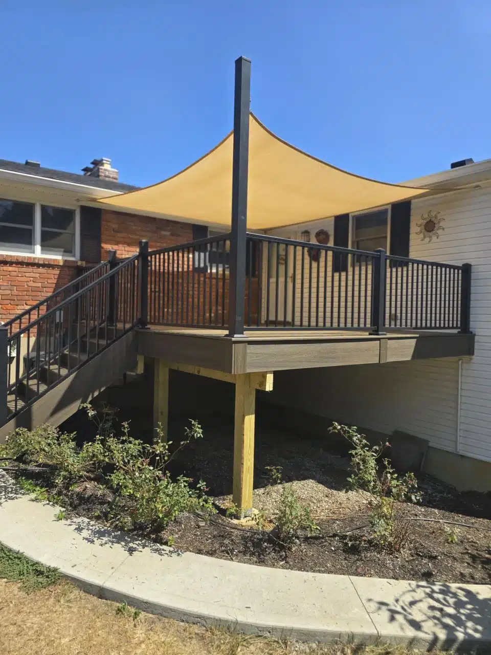 Side view of the elevated deck with black aluminum railing and a shade sail above. The deck is supported by posts with a landscaped area around the base