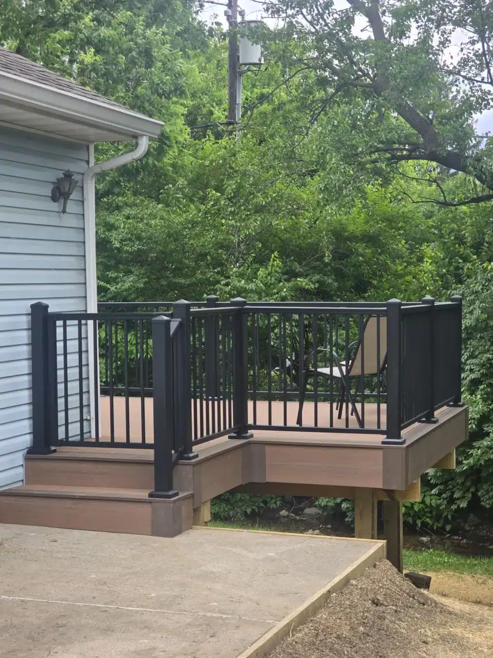 Close-up view of the deck stairs, showing black aluminum railing and composite decking. The deck leads off from the side of the house, with a chair visible on the deck and greenery in the background.