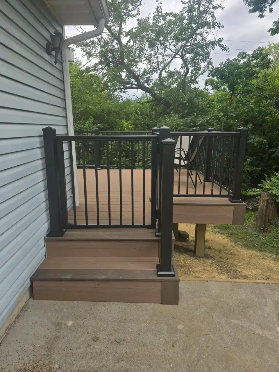 Corner view of the deck, showing the black aluminum railing, composite decking, and a chair on the deck. The house's blue siding and green trees are visible in the background.