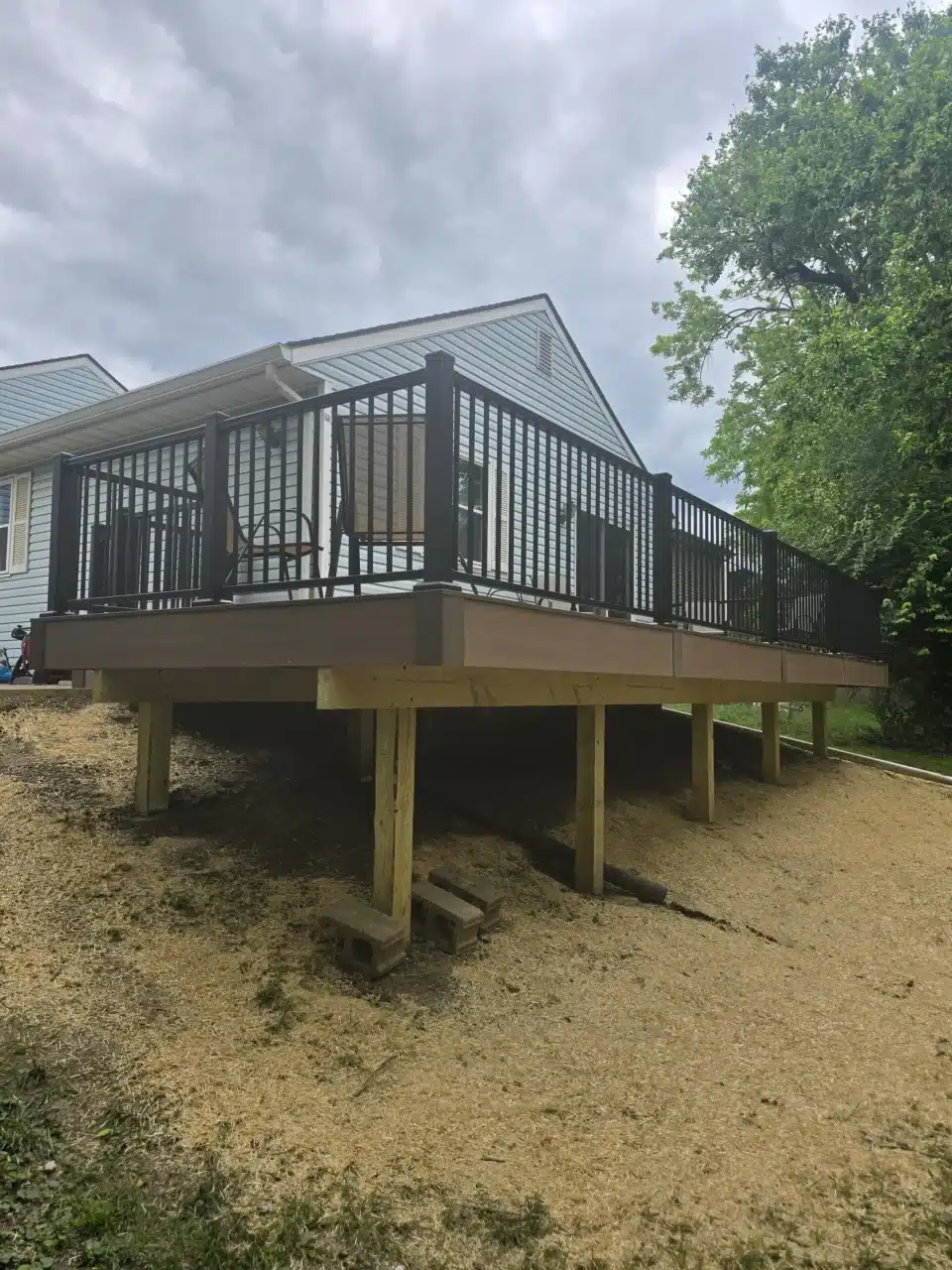 Side view of the deck, showing the black aluminum railing, the understructure supported by posts, and the back of the house with trees in the background