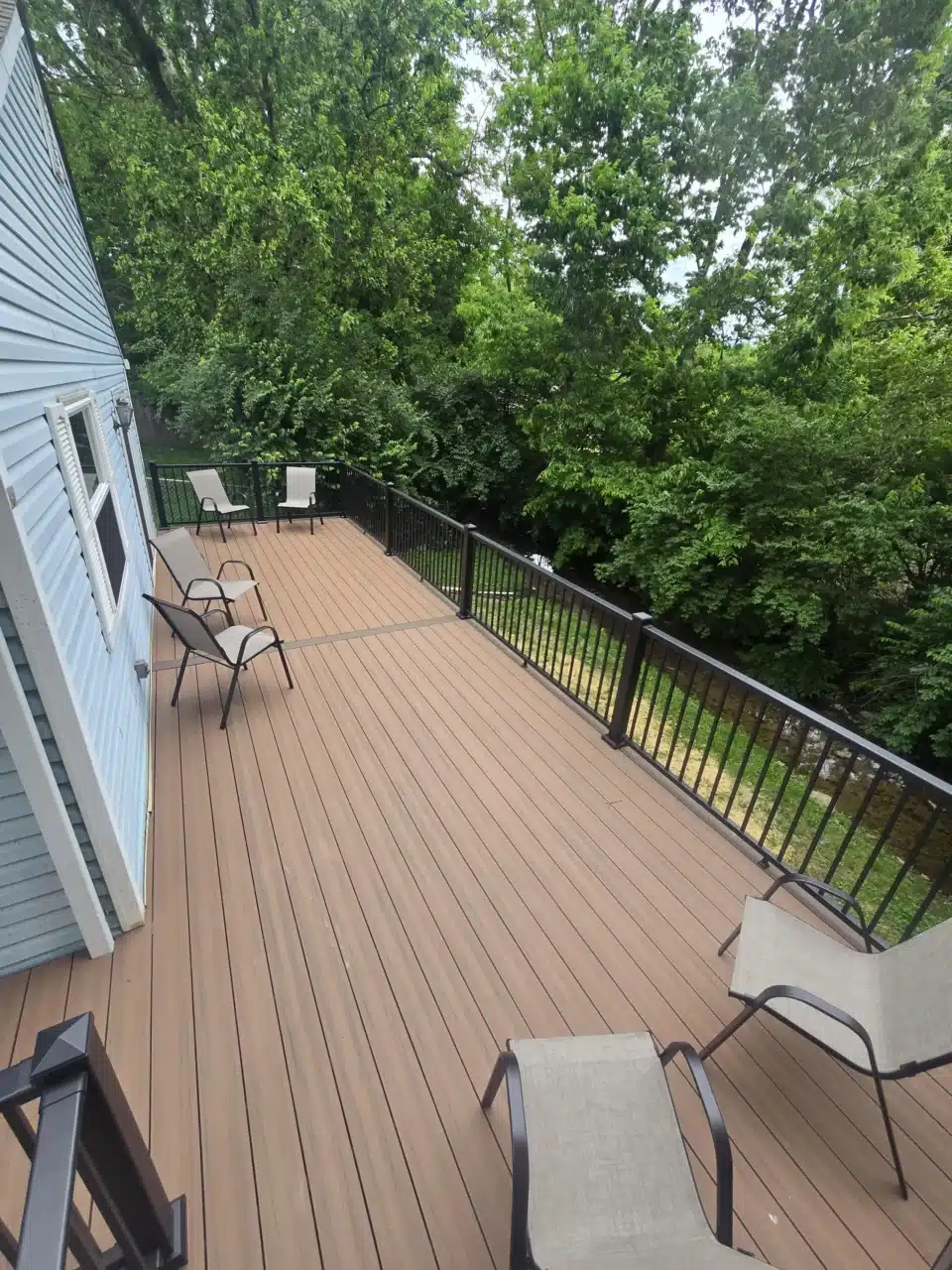 Wide-angle view of the deck, showing composite decking and black aluminum railing. Two chairs are placed on the deck, with green trees in the background.
