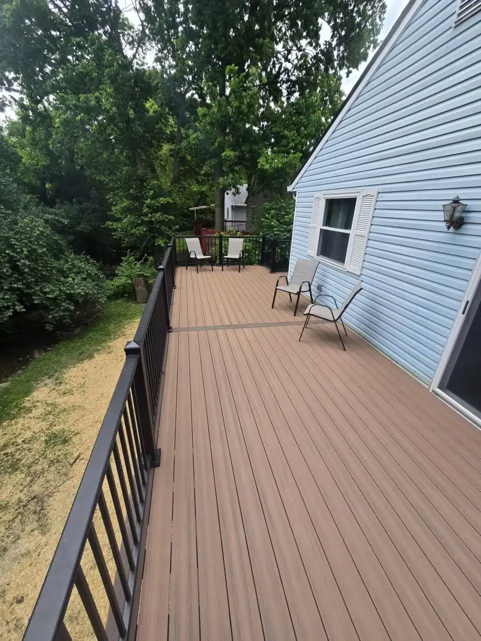 View of the deck from above, showing composite decking, black aluminum railing, and two chairs. The back of the house with blue siding and green trees in the background.