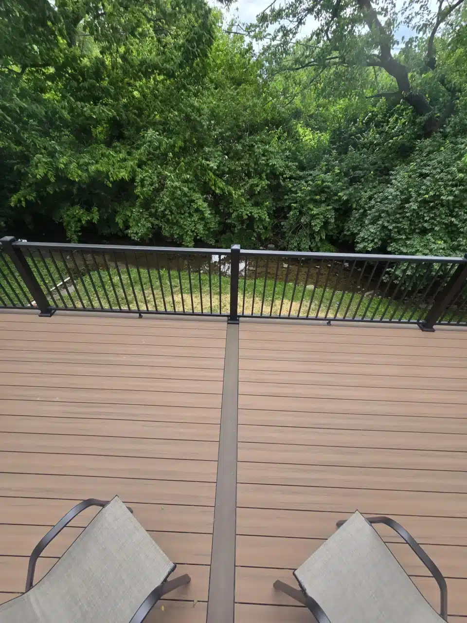 View from the deck, showing composite decking, two chairs, and black aluminum railing. The background features greenery and trees.