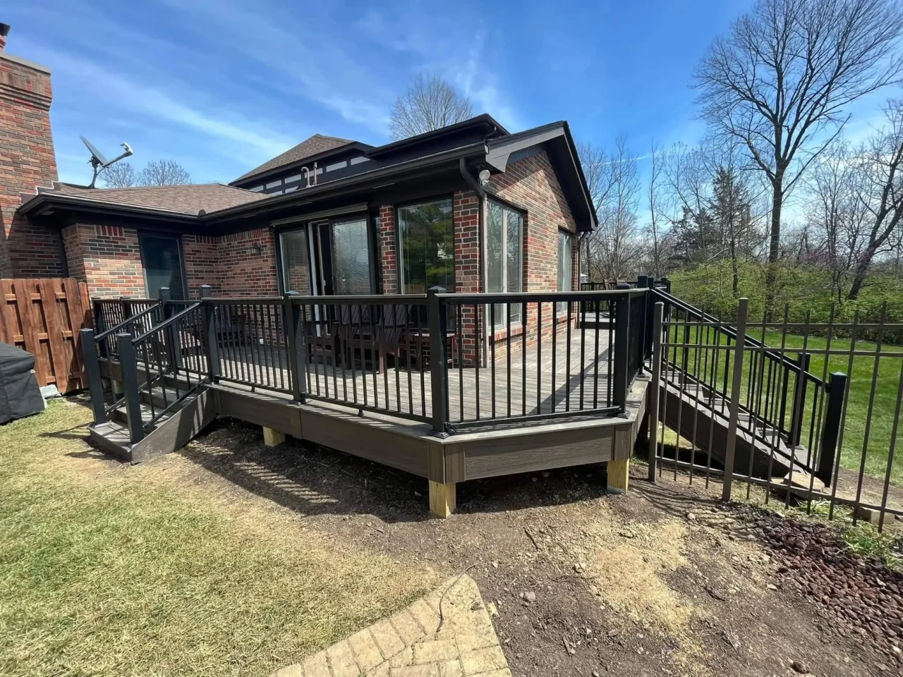 Side view of a raised deck with black railing, attached to a brick house. A staircase leads down to the yard, with trees and grass visible in the background.