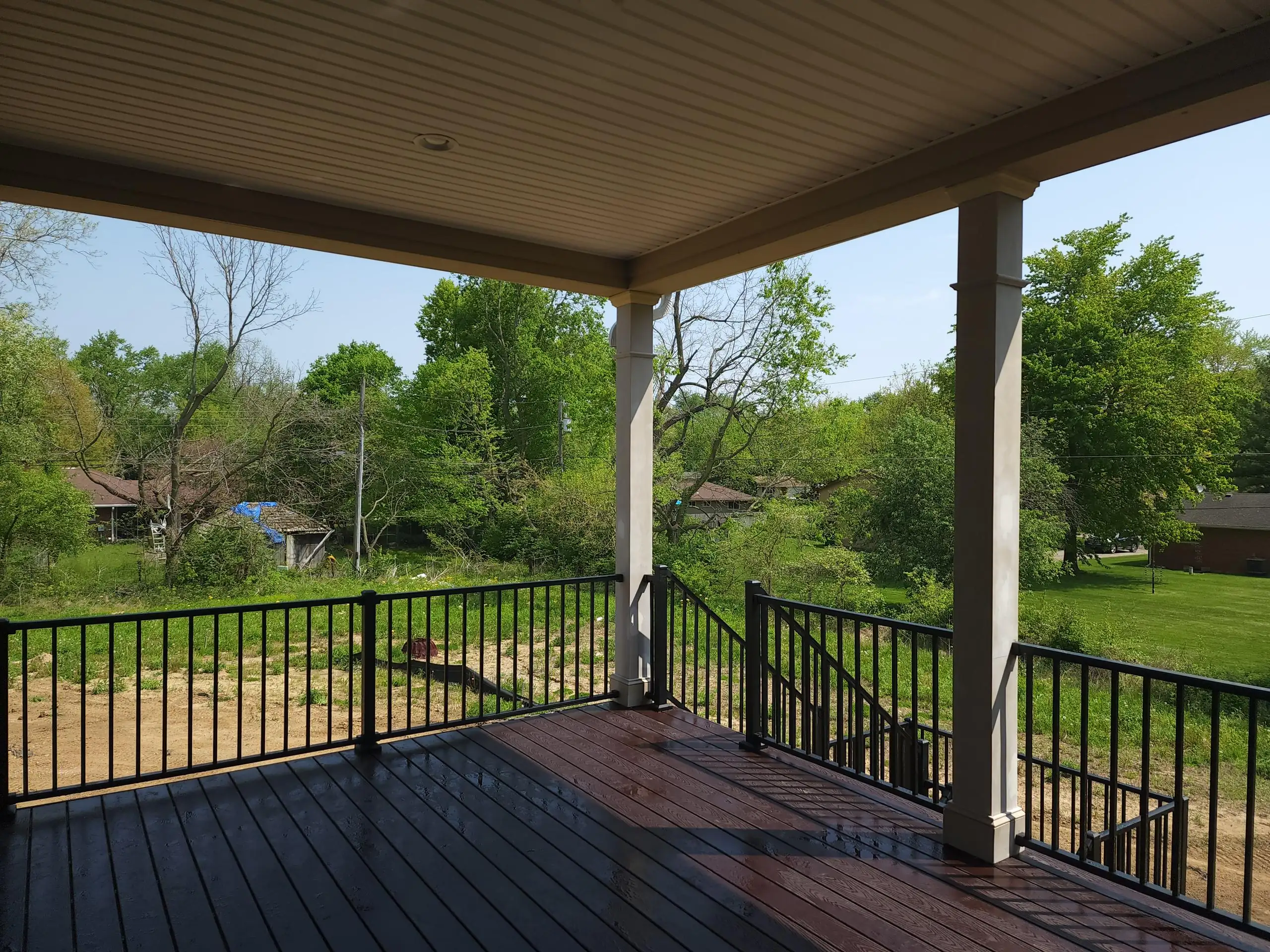 Best Composite Deck Boards A covered outdoor deck featuring dark composite decking boards, black aluminum railings, and two light beige support columns. The deck overlooks a grassy backyard with scattered trees and residential homes visible in the background under a bright, clear sky.