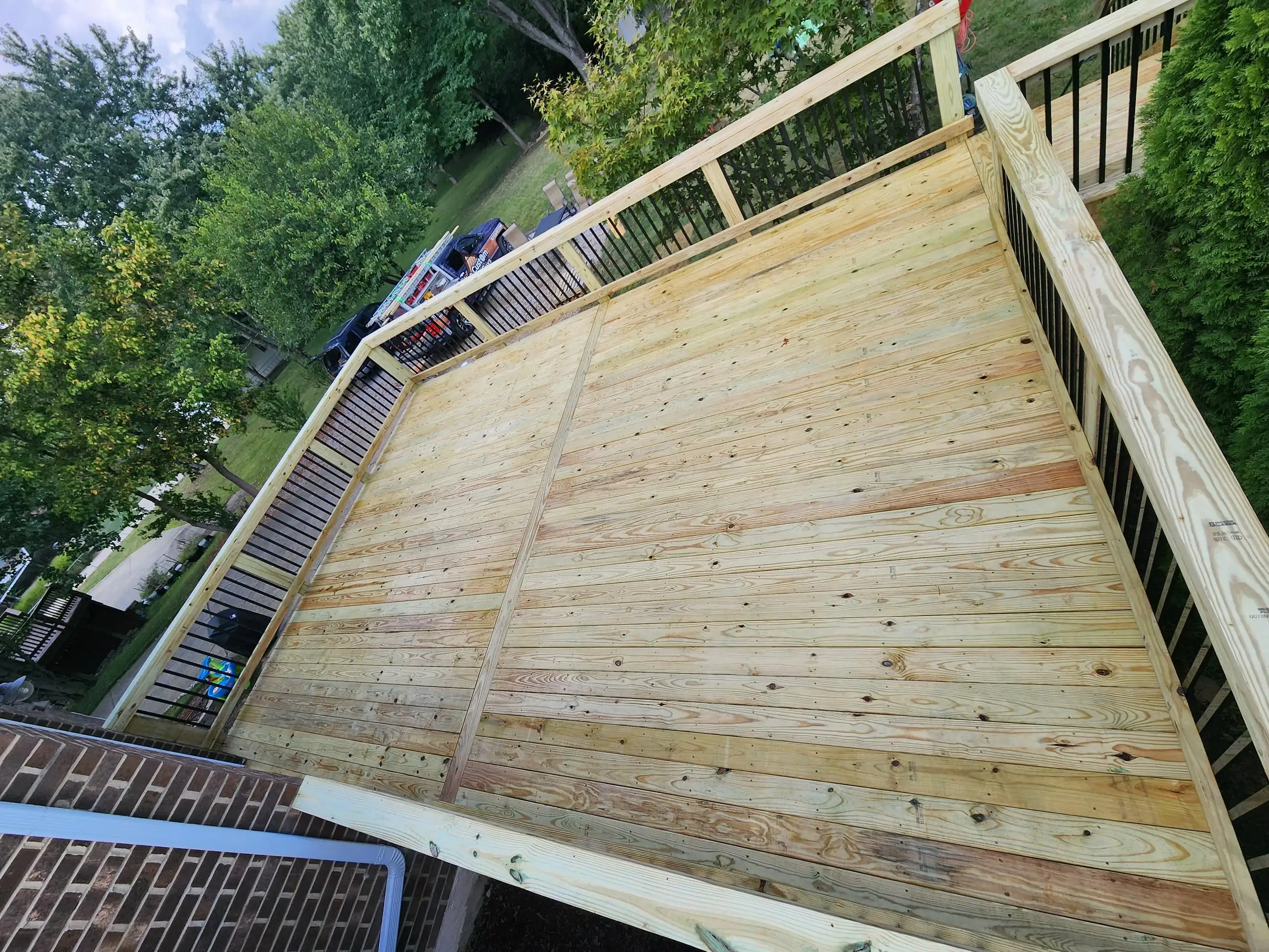 A newly constructed outdoor wooden deck. The deck has a light wood finish and features black railings with wooden posts. In the background, there are trees, some greenery, and what appears to be a vehicle.