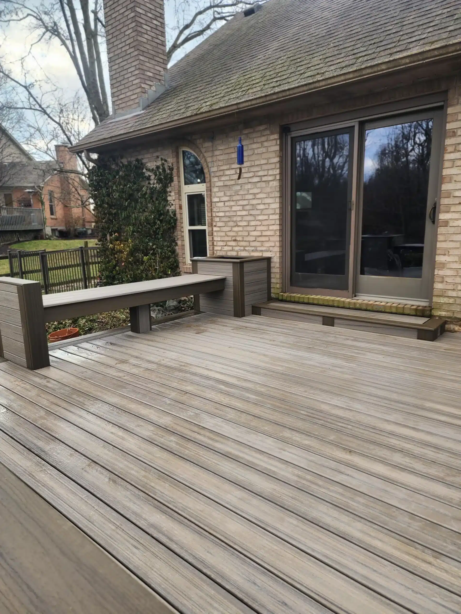 Composite Decking Brands A residential composite deck featuring wide, light gray composite deck boards with a multi-tonal, weathered-wood look. The deck includes a built-in bench and a small planter box, both constructed from the same composite material, situated against a brick house with large sliding glass doors.
