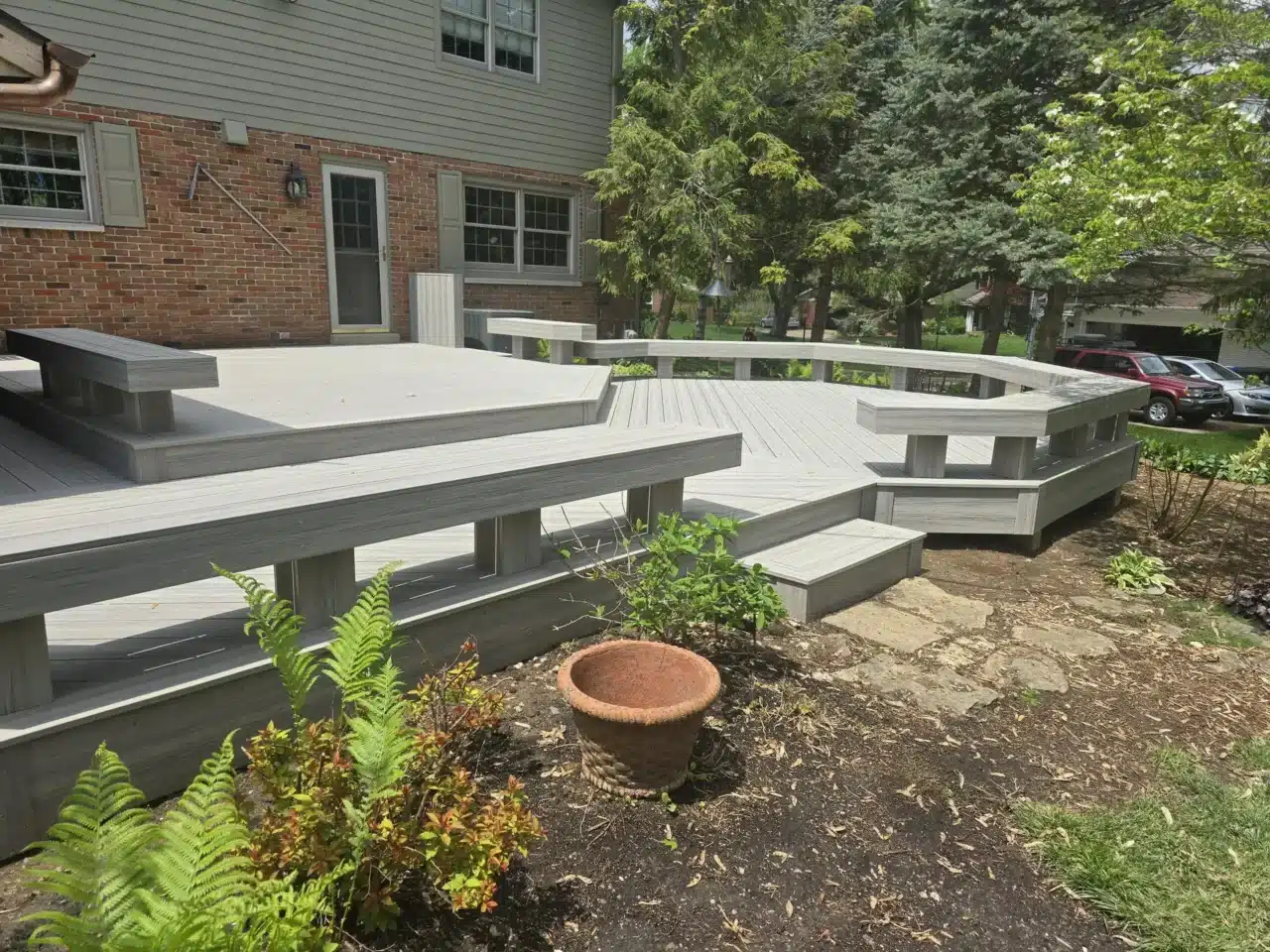An elevated outdoor deck made of light gray composite material. The deck features a multi-level design with different sections connected by steps. Integrated bench seating with backs is built into the edges of the deck in several places.