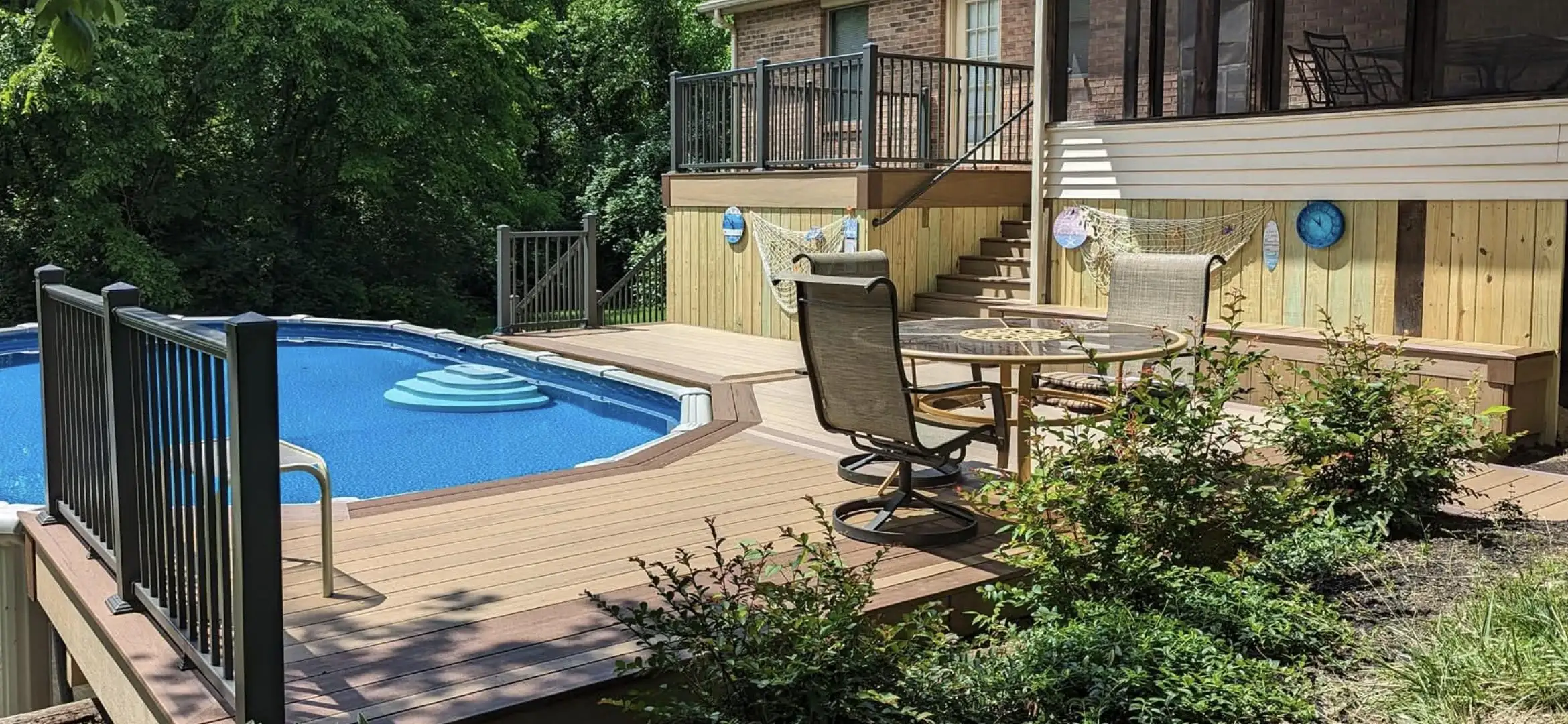 wood deck cost per sf An elevated outdoor living space with a large above-ground swimming pool and a multi-level deck constructed with composite decking. The pool has blue water and stairs leading into it. The main deck area features a round table with two swivel chairs and is decorated in a nautical theme, with fishing nets and blue wall clocks on the house's wooden siding.