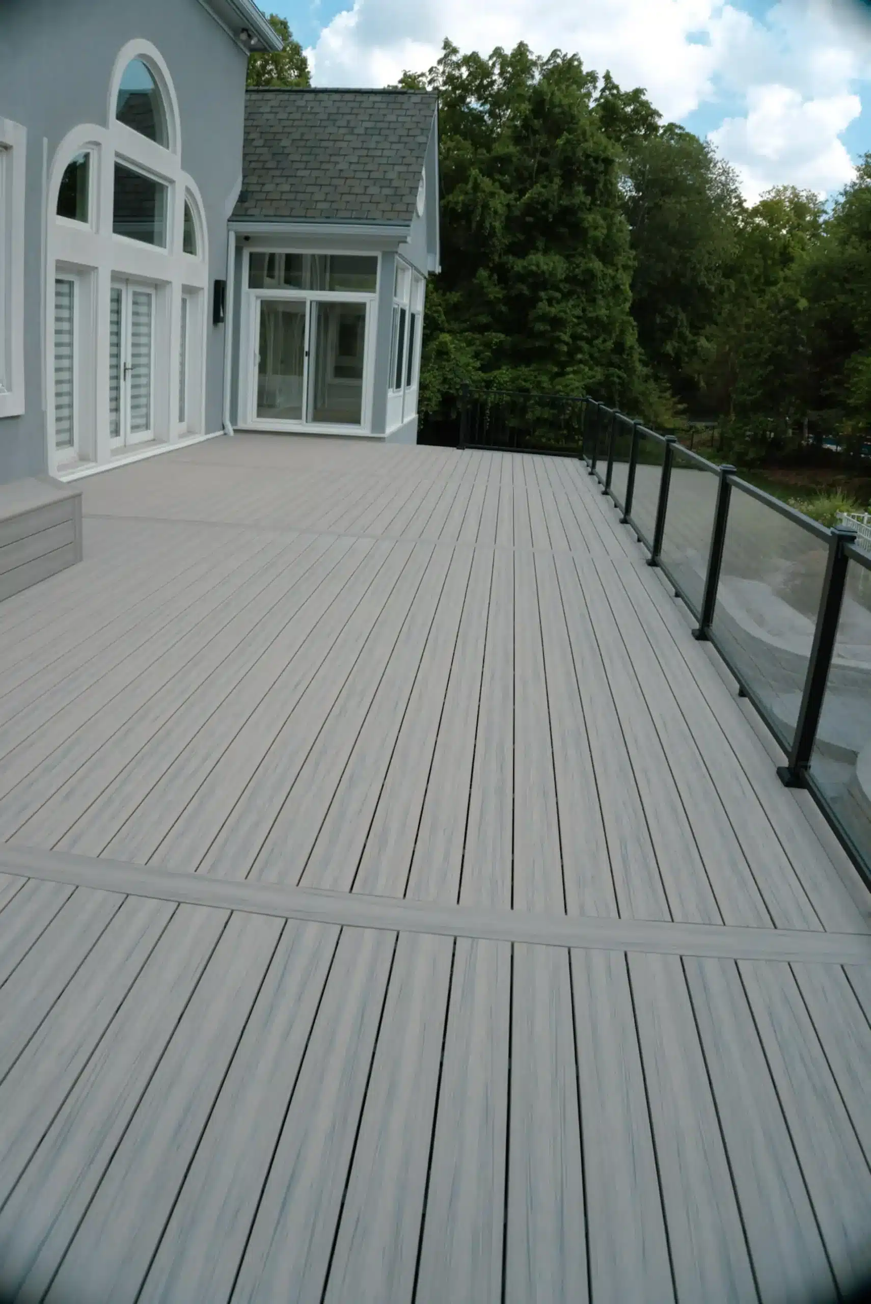A large, modern grey composite deck with black and glass railings surrounding the perimeter. The deck is attached to the back of a light grey house and overlooks a wooded area.