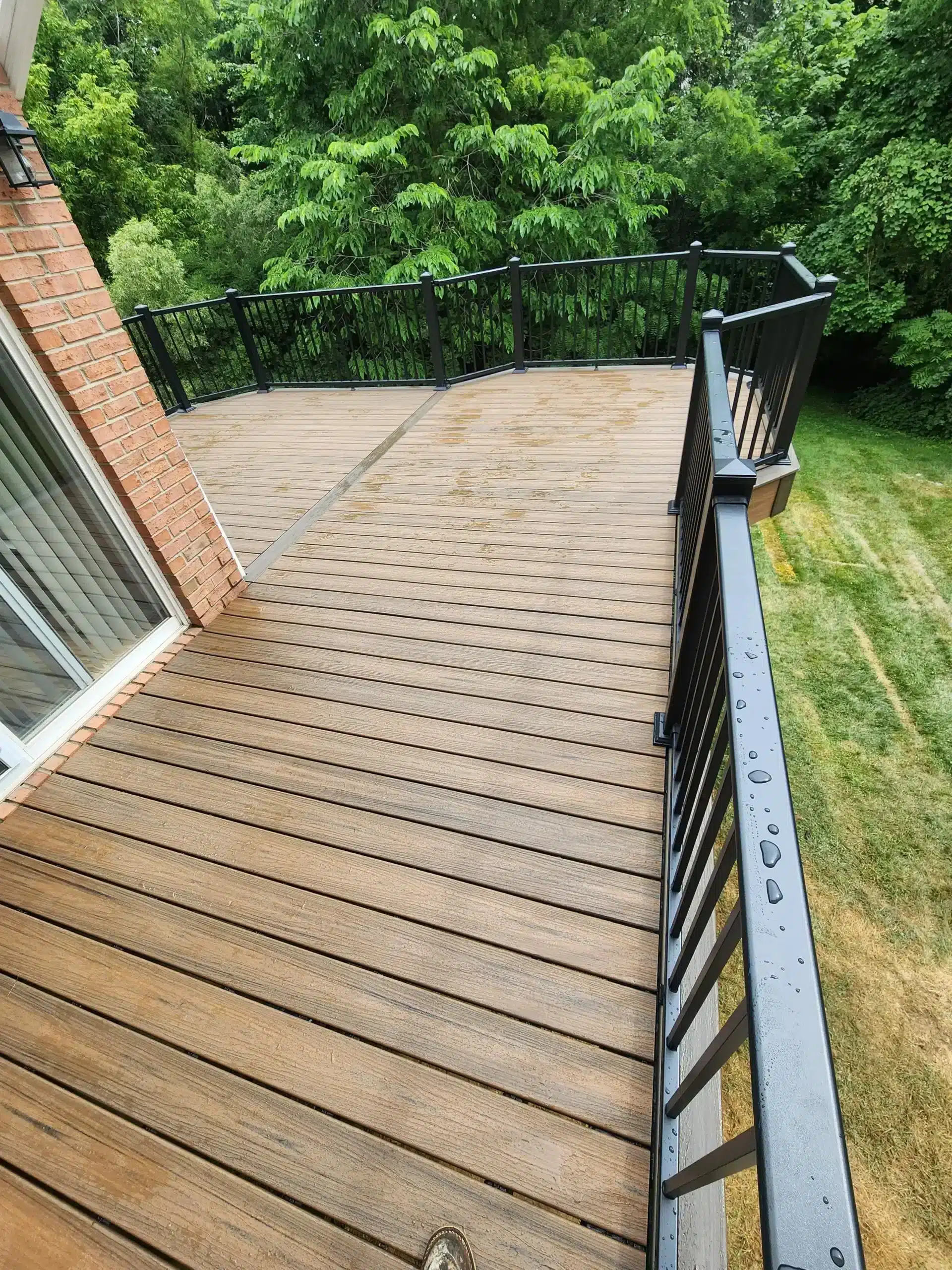 how long does trex decking last A high-angle view of a new brown composite deck with black metal railings, attached to a house with a brick exterior. The deck boards are wet, and the railing curves around the deck's edge, which overlooks a lush green yard and trees.